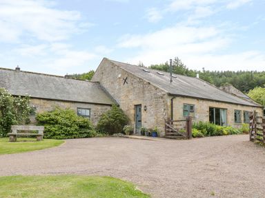 A stone house with a gravel driveway green bushes and a wooden bench at The Steadings in Rothbury