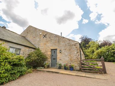 A stone building with a gray door and flower pots on the ground next to a wooden gate and gravel driveway at The Steadings in Rothbury