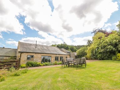A stone house with a sloped roof next to a wooden table and chairs on a lawn at The Steadings in Rothbury