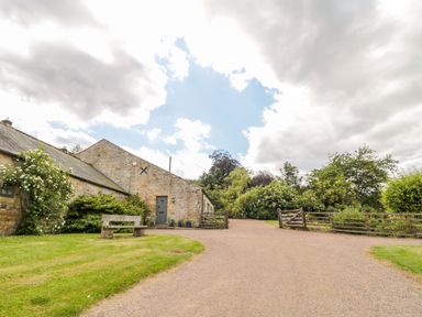 A stone building with a wooden door and bench outside next to a gravel driveway and wooden gates at The Steadings in Rothbury