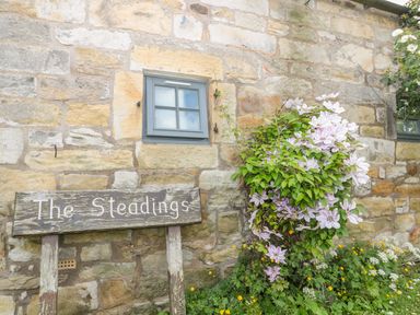 A stone wall with a small window a wooden sign saying The Steadings and flowering plants at The Steadings in Rothbury