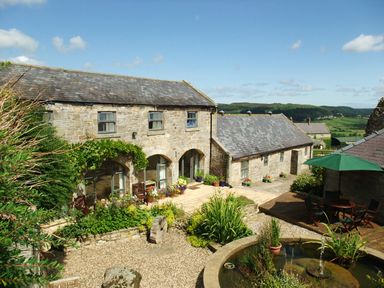 A stone building with arched windows a patio garden and a round pond with a fountain at The Steadings in Rothbury