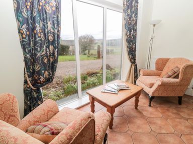 A sitting area with two armchairs a wooden coffee table with magazines and large windows with patterned curtains at The Steadings in Rothbury