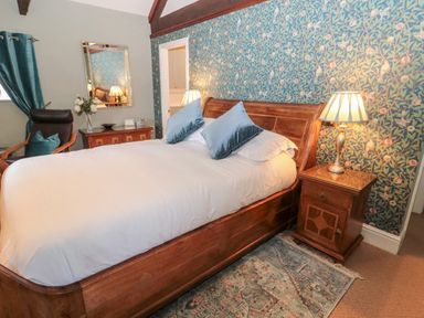 A bedroom with a wooden bed with white bedding and blue pillows beside a wooden nightstand with a lamp and floral wallpaper at The Steadings in Rothbury
