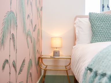 A bedside table with a lamp next to a bed in a bedroom with pink and green wallpaper at See Two Seas Cape Cornwall in St Just