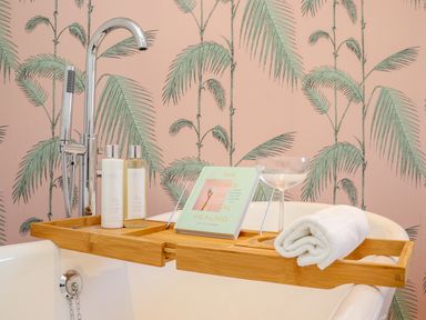 A bathtub with a wooden tray holding two bottles, a book, a glass, and a rolled towel at See Two Seas Cape Cornwall in St Just