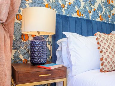 A bedside table with a blue lamp and a book next to a bed with white pillows and a patterned cushion at See Two Seas Cape Cornwall in St Just