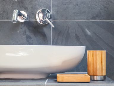 A bathroom sink with chrome faucet and wooden soap dispenser and tray on a gray tiled wall at See Two Seas Cape Cornwall in St Just