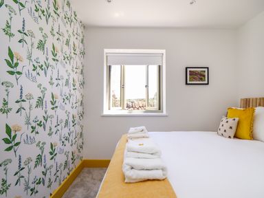 A bedroom with a bed towels on yellow blanket floral wallpaper window and framed picture at See Two Seas Cape Cornwall in St Just