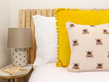 A close-up of a bed with white sheets yellow and bee-patterned cushions and a bedside table with a bee-patterned lamp and a book at See Two Seas Cape Cornwall in St Just