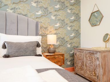A bedroom with a bed a bedside table with a lamp a wooden cabinet a hexagonal wall mirror and patterned wallpaper at See Two Seas Cape Cornwall in St Just
