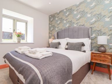 A bedroom with a bed with gray and white bedding floral wallpaper window with flowers on the sill and wooden bedside table with lamp at See Two Seas Cape Cornwall in St Just