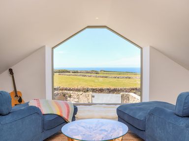 A sitting area with blue armchairs a round glass table and a guitar by a large window overlooking fields and sea at See Two Seas Cape Cornwall in St Just