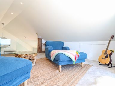 A room with blue upholstered furniture a glass coffee table on a woven rug an acoustic guitar on a stand and a sloped ceiling at See Two Seas Cape Cornwall in St Just