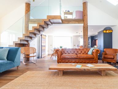 A living room with a leather sofa blue armchair wooden coffee table and a staircase with glass railing at See Two Seas Cape Cornwall in St Just