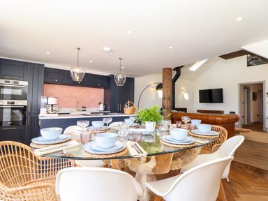 A dining area with a glass table set with dishes and glasses next to a kitchen with dark cabinets and pink backsplash at See Two Seas Cape Cornwall in St Just