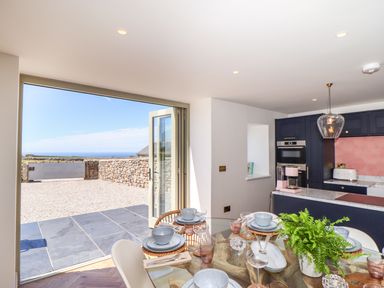 A kitchen and dining area with glass table set for a meal and open doors to an outdoor patio with stone walls at See Two Seas Cape Cornwall in St Just