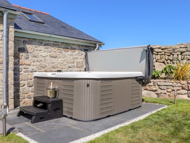 An outdoor hot tub with steps and a champagne bucket on a tiled patio next to a stone house and garden at See Two Seas Cape Cornwall in St Just