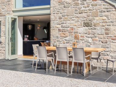 An outdoor dining area with a wooden table and eight chairs on a tiled patio against a stone wall at See Two Seas Cape Cornwall in St Just
