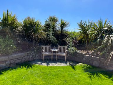 A garden seating area with two wooden chairs and a small table surrounded by plants and stone walls at See Two Seas Cape Cornwall in St Just