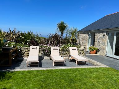 Three beige lounge chairs on a stone patio next to a stone house with plants and green grass at See Two Seas Cape Cornwall in St Just