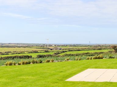 A green field with shrubs and a paved area overlooking farmland with wind turbines and buildings at Pen Tewan in Sennen