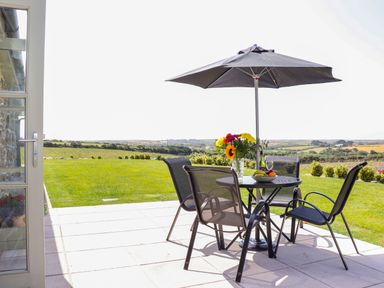 An outdoor patio with a round table and four chairs under a large umbrella with flowers fruit and wine on the table at Pen Tewan in Sennen