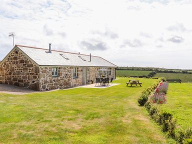 A stone house with a patio table and chairs and a picnic bench on a lawn with flower beds and fields in the background at Pen Tewan in Sennen