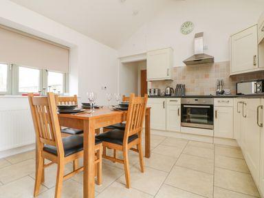 A kitchen with a wooden dining table and chairs set with plates and wine glasses next to cabinets and appliances at Pen Tewan in Sennen