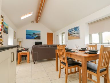 A dining area with a wooden table and four chairs set with plates and glasses next to a living area with two brown sofas a side table a lamp paintings on the walls and a skylight at Pen Tewan in Sennen