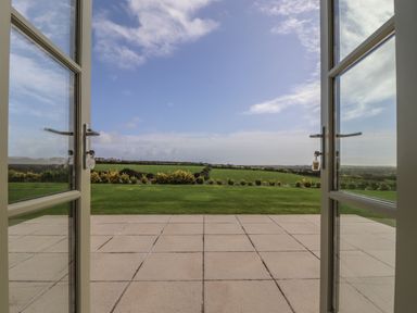 An open patio door leading to a tiled patio and a lawn with fields and a partly cloudy sky at An Gwel in Sennen
