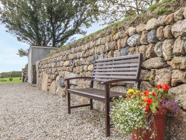 A wooden bench beside a stone wall with a planter of flowers on a gravel path at An Gwel in Sennen