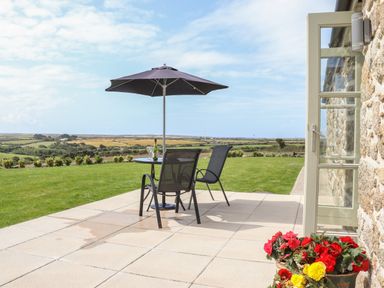 A patio with two chairs a table and an umbrella overlooking fields with flowers and a stone wall at An Gwel in Sennen