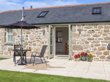 A stone exterior house with an open double door leading inside a patio table with an umbrella and two chairs and flower pots on the ground at An Gwel in Sennen