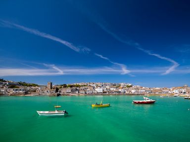 A harbor with boats on turquoise water and a town with buildings and a church in the background at An Gwel in Sennen