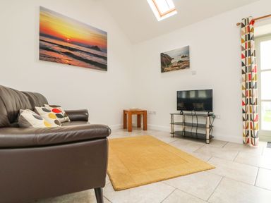 A living room with a brown leather sofa, colorful cushions, a television on a stand, a small wooden table, wall art, a skylight window, and patterned curtains at An Gwel in Sennen