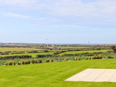 A grassy garden with a paved area overlooking farmland and wind turbines at An Garth in Sennen