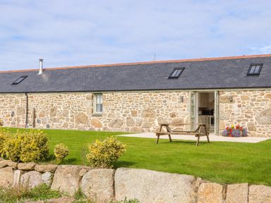 A stone building with a slate roof and skylights in a green garden with a wooden bench and flower pots at An Garth in Sennen