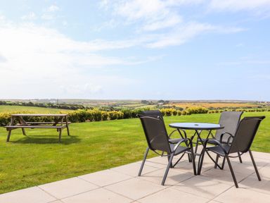 A patio with a round glass table and four chairs and a grassy lawn with a wooden picnic bench at An Garth in Sennen