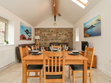 A dining area with a wooden table set for four and a living area with two sofas and a TV at An Garth in Sennen