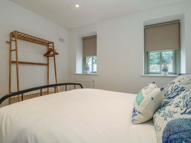 A bedroom with a bed with pillows a wooden clothes rack and two windows with blinds at An Garth in Sennen
