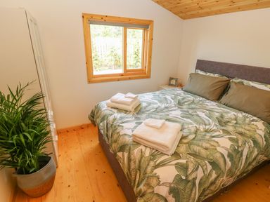 A bedroom with a bed with leafy patterned bedding white towels on the bed a window wooden floor and ceiling a plant in a pot and a white wardrobe at Willow Lodge in St Florence