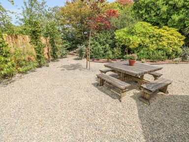 A garden area with a wooden picnic table and benches on gravel surrounded by trees and shrubs at Willow Lodge in St Florence
