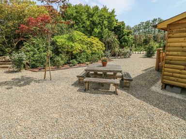 An outdoor area with a wooden picnic table and benches on gravel near a wooden building and surrounded by trees and shrubs at Willow Lodge in St Florence