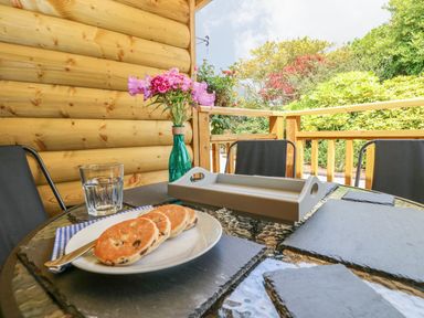 A table with a plate of three scones a glass of water a vase with flowers and patio chairs on a wooden deck outside at Willow Lodge in St Florence