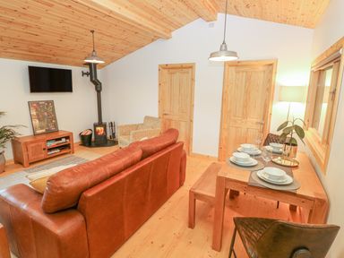 A living room with a brown leather sofa, a wood burning stove, a wall mounted TV, and a dining table set for four at Willow Lodge in St Florence