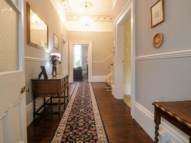 A hallway with wooden floor a patterned runner rug a wooden side table with flowers a mirror and a chandelier at Holcombe House in Torquay