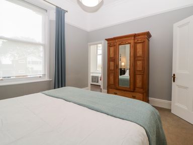 A bedroom with a bed covered with a blanket a wooden wardrobe with a mirror and a window with curtains at Holcombe House in Torquay