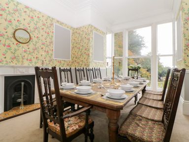 A dining room with a long wooden table set with bowls wine glasses and a brass candelabra surrounded by wooden chairs and floral wallpaper at Holcombe House in Torquay