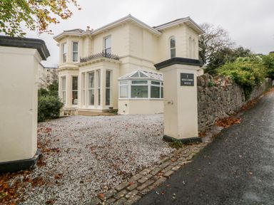 An exterior view of a two-story cream house with large windows a gravel driveway and stone wall at Holcombe House Torquay